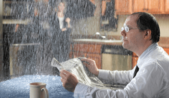 Professional man in business attire wearing glasses drinking coffee sitting at dining table at home reading newspaper ignoring rain and leaking roof leak in house with woman wearing all black in background holding umbrella.
